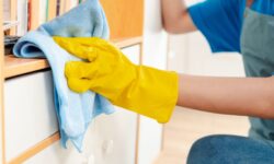 "A professional cleaner wearing yellow gloves and wiping a shelf with a blue microfiber cloth."