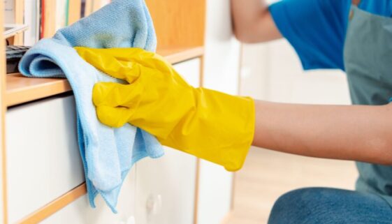 "A professional cleaner wearing yellow gloves and wiping a shelf with a blue microfiber cloth."